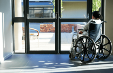A woman sitting in a wheelchair looks out glass doors in a nursing home