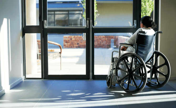 A woman sitting in a wheelchair looks out glass doors in a nursing home