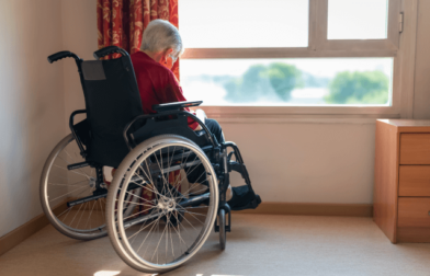 An older adult in a wheelchair sits alone by a sunny window in a nursing home room, looking down with their back turned, suggesting isolation or neglect