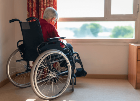 An older adult in a wheelchair sits alone by a sunny window in a nursing home room, looking down with their back turned, suggesting isolation or neglect